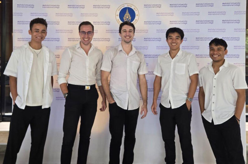 Oswaldo Medellin with classmates standing in front of a Mahidol University backdrop, all wearing white school shirts and dark pants on their last day of classes.