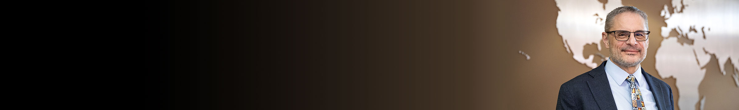 Director of the International Business Institute Jeff Joireman standing in front of a metallic world map display, wearing a suit and patterned tie, with a dark gradient background to the left.