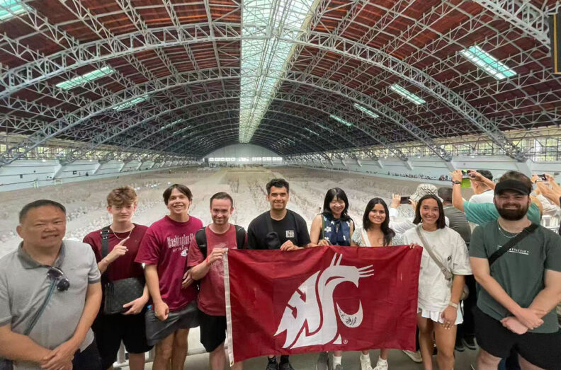 A group of Carson College students stand inside a large industrial hall of Terra Cotta Warriors, holding a crimson Washington State University flag during a study abroad visit.