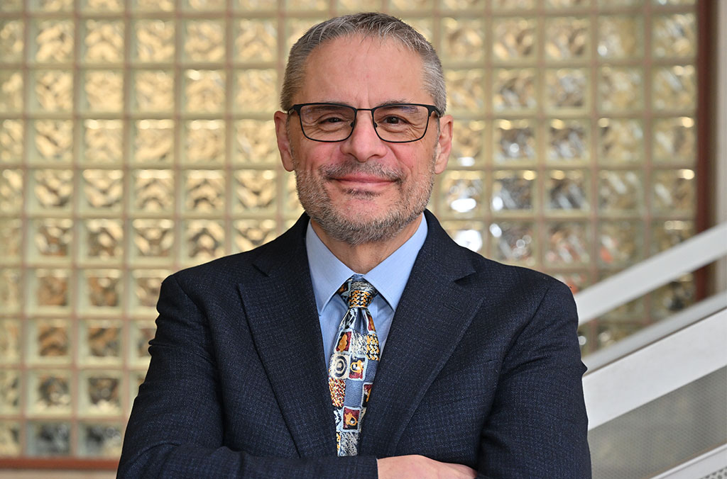 International Business Institute Director Jeff Joireman stands with arms crossed in a suit and patterned tie inside a campus building with a glass‑block wall behind him.
