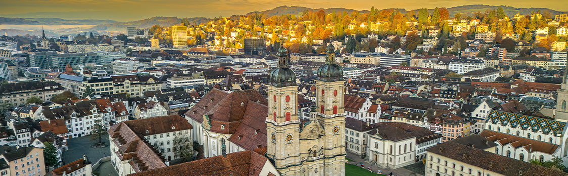 Wide aerial view of St. Gallen, Switzerland, highlighting the Abbey of St. Gall with twin cathedral towers, surrounded by red‑roofed buildings and autumn hills.