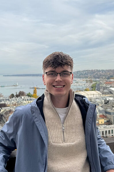 Stan de Keijzer at a scenic overlook above a Swiss lakeside city, with clustered rooftops, cranes, and the shoreline below.