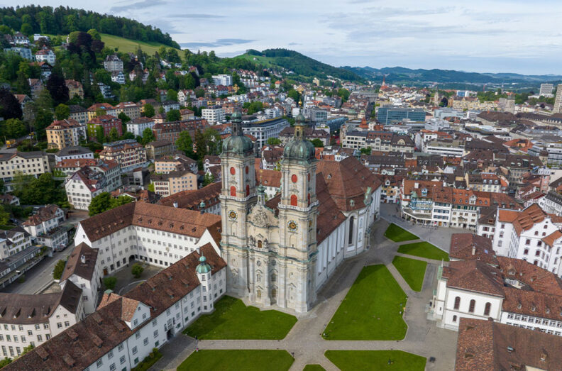 Aerial view of a historic European city centered on a large cathedral with twin towers, surrounded by red‑roofed buildings, green hillsides, and winding streets.