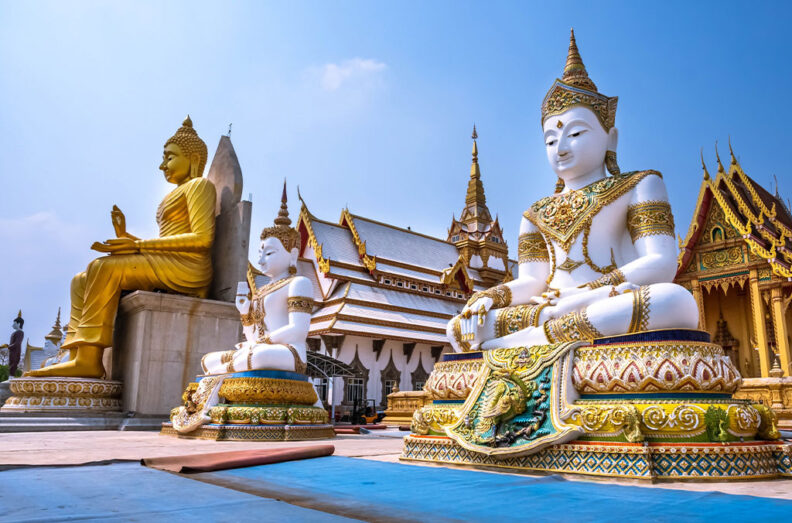 Ornate Buddhist statues sit in front of a richly decorated Thai temple, with gold and white figures arranged across a sunlit courtyard.