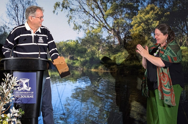 Joseph Gladstone stands at a University of Melbourne podium while Michelle Evans applauds beside a river backdrop.