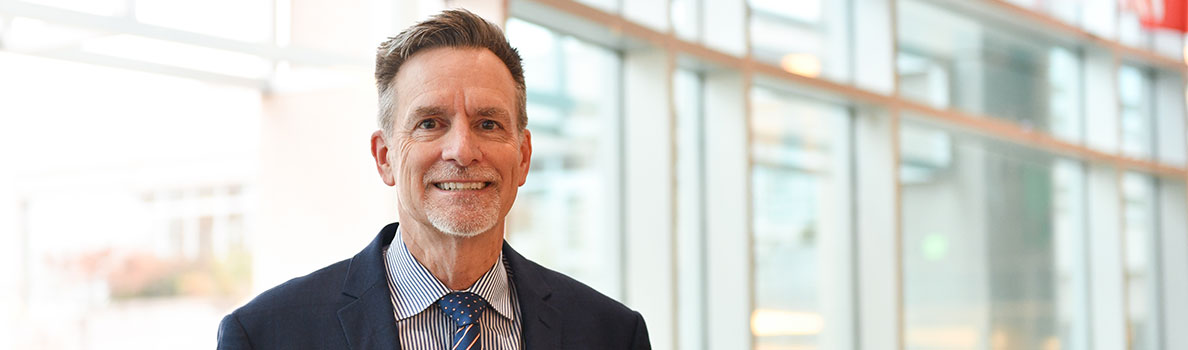 Bob Harrington wearing a dark suit with a striped shirt and patterned tie, standing in a bright indoor space with large glass windows in the background.