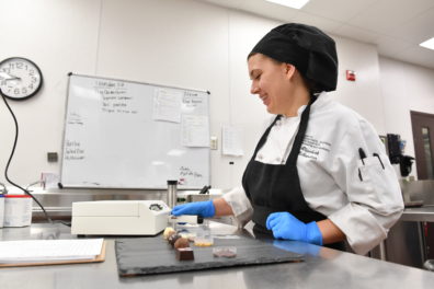 Elizabeth Nalbandian in a commercial kitchen wearing a chef’s uniform and blue gloves, working at a counter with an AquaLab moisture analyzer and assorted chocolate pieces on a black slate.