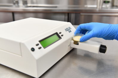 Gloved hand placing a petri dish into an AquaLab moisture analyzer on a stainless steel countertop in a laboratory setting.