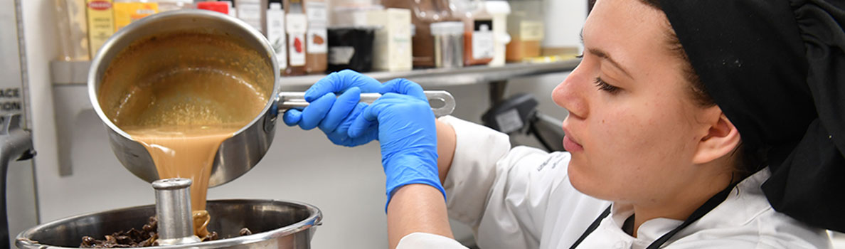Elizabeth Nalbandian wearing a chef’s uniform and blue gloves, pouring a caramel-colored mixture from a saucepan into a metal bowl in a commercial kitchen.