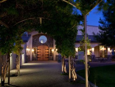 Arched walkway covered with greenery leading to a large wooden door of a stone building, illuminated by warm lights at dusk.