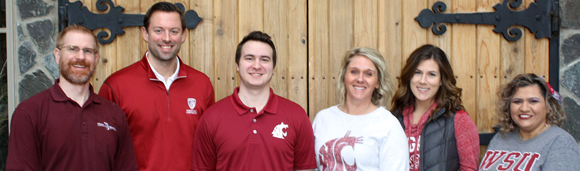 Group of six people standing in front of large wooden doors with decorative black iron hinges, wearing Washington State University apparel in shades of crimson, gray, and white.