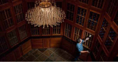 Person selecting a wine bottle from wooden racks in a dimly lit wine cellar with a large crystal chandelier overhead.