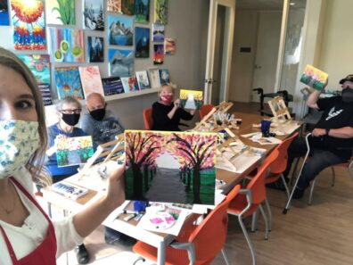 Art class participants seated at a long table displaying their paintings of a tree-lined path with pink blossoms, while a person in the foreground holds up a similar finished painting. Colorful artwork decorates the walls in the background.