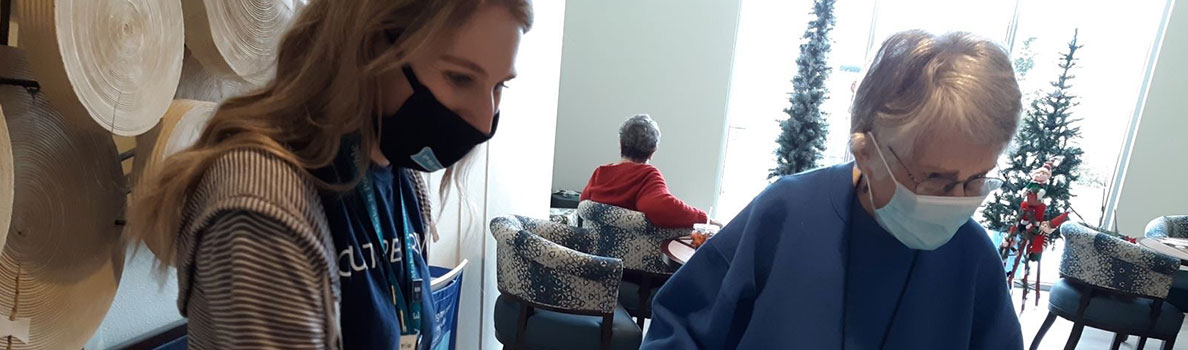 Two people engaged in an indoor activity at a table, with decorative circular wall art on the left and holiday trees in the background. Another person is seated further back wearing a red top.