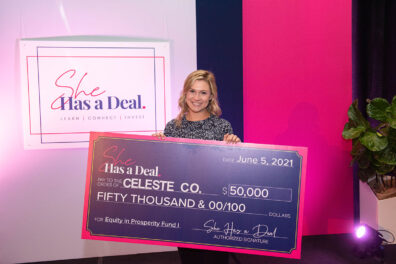 Emma Claire Spring holding an oversized ceremonial check for $50,000 from the She Has a Deal hotel pitch competition, standing in front of a display with the event logo.