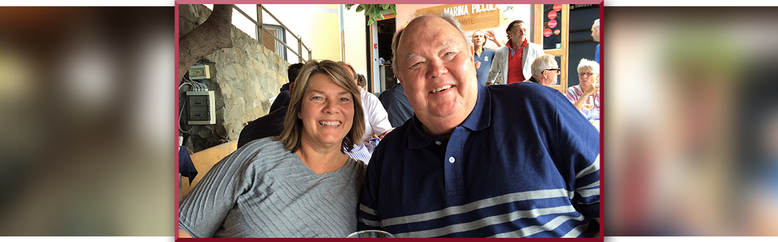 Two people seated at an outdoor restaurant table with a glass in the foreground; Larry Culver is on the right wearing a dark shirt with light horizontal stripes.