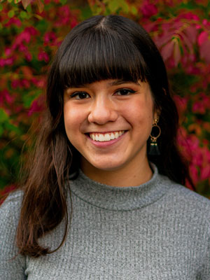 Portrait of Makenna Price wearing a gray ribbed turtleneck sweater, standing outdoors in front of vibrant red and green foliage.
