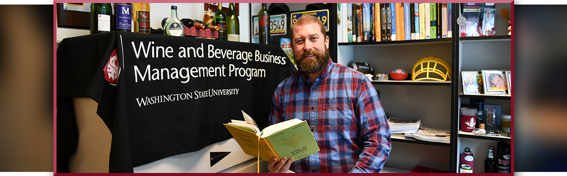 Byron Marlowe holding an open book in an office with shelves of books and memorabilia, next to a table draped with a Washington State University Wine and Beverage Business Management Program banner.
