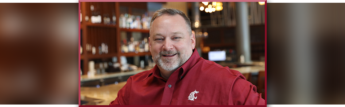Anthony Anton wearing a crimson WSU-branded shirt, seated in a bar or restaurant setting with shelves of bottles in the background.