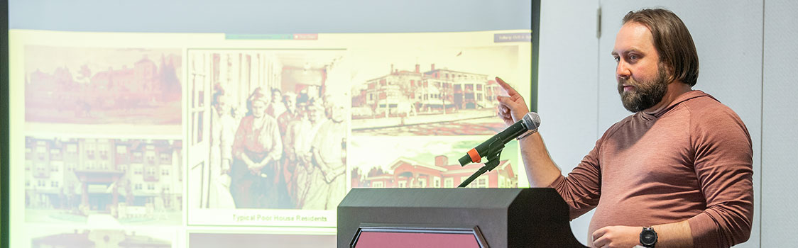 Murry Mercier presenting at a Washington State University podium during a lecture on senior living, with a projected slide showing historical images and the text “Where did Senior…” partially visible.