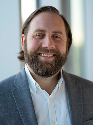 Murry Mercier wearing a light gray suit jacket over a white collared shirt, standing in front of large windows with natural light.