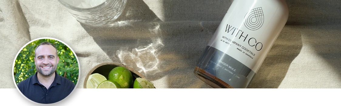WithCo Cocktails COO Shaun McKinley pictured alongside a styled product photo featuring a bottle of WithCo cocktail mixer labeled “Ginger Mule,” a glass with ice, and a bowl of fresh limes on a light fabric surface in natural sunlight.