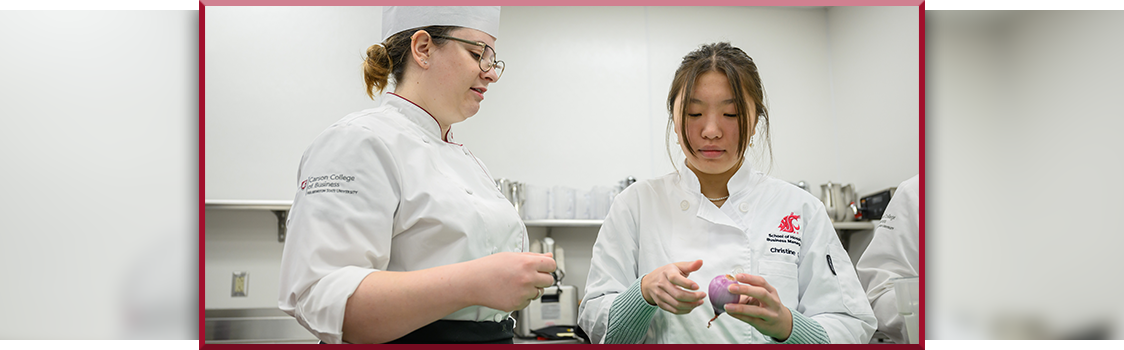 Jessica Murray, wearing a chef’s uniform and hat, standing in a commercial kitchen and talking with another culinary student who is holding a small purple vegetable.