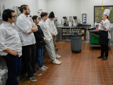 Jessica Murray, wearing a chef’s hat and uniform, speaks to a group of culinary students standing in a commercial kitchen with stainless steel counters and equipment.