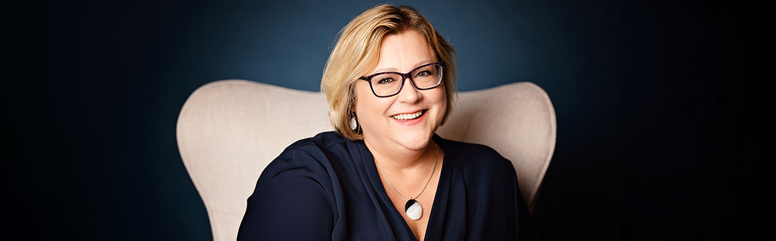 April Reddout seated in a light-colored chair against a dark blue background, wearing a dark top and a pendant necklace.