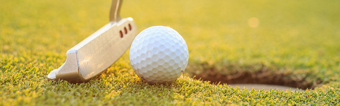 Close-up of a golf ball positioned near a hole on a green, with a putter aligned behind the ball under warm sunlight.