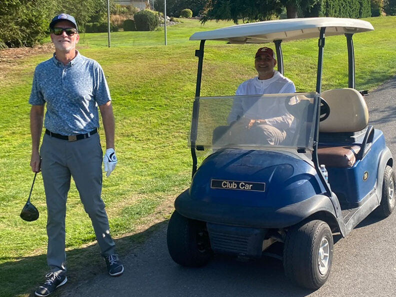 Golf cart parked on a paved path with one person seated inside and another standing nearby holding a golf club on a grassy area during the SHBM First Annual Golf Tournament.