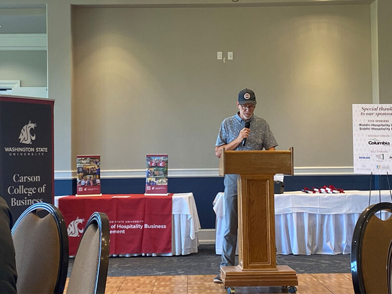 Bob Harrington speaking at a podium during an event, with Washington State University Carson College of Business and School of Hospitality Business Management banners and tables in the background.