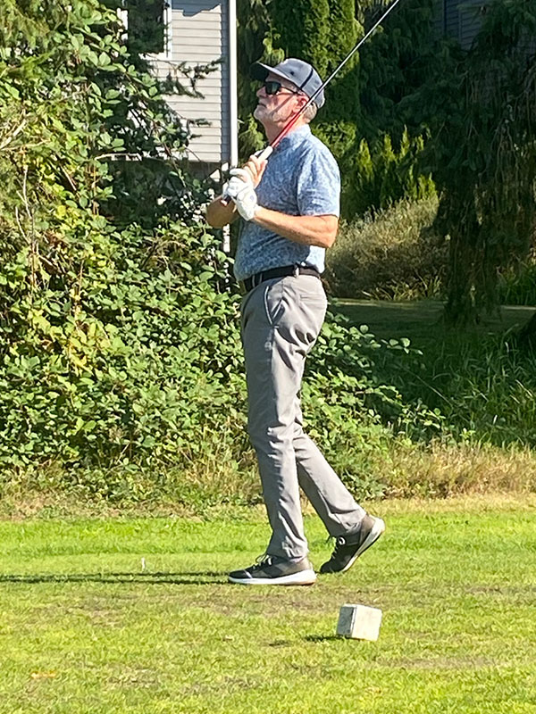 Bob Harrington standing on a golf course holding a club after a swing, wearing a patterned polo shirt, gray pants, golf gloves, and sneakers, with greenery and a building in the background.