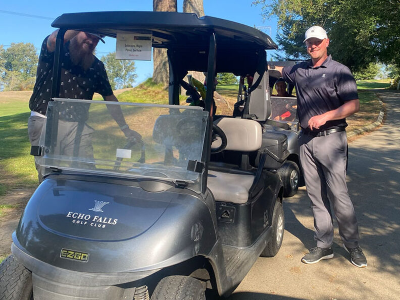 Two golf carts parked on a paved path at Echo Falls Golf Club during the SHBM First Annual Golf Tournament, with two participants standing nearby.
