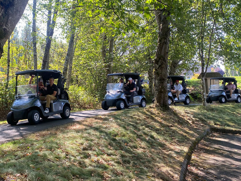 Four golf carts lined up on a shaded path surrounded by trees during the SHBM First Annual Golf Tournament.