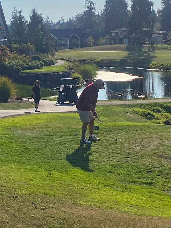 Golfer preparing to take a shot on a grassy area near a pond, with a golf cart and another person visible in the background during the SHBM First Annual Golf Tournament.