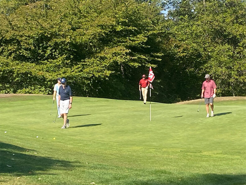 Four golfers on a putting green near a red flagstick, with golf balls scattered around and dense green trees in the background during the SHBM First Annual Golf Tournament.