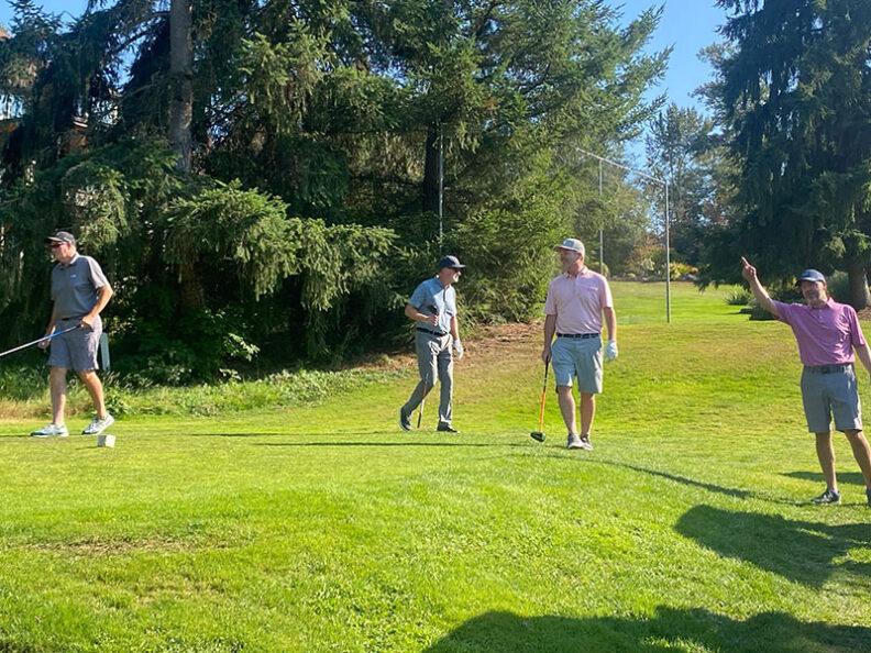 Four golfers standing on a green tee area with clubs, surrounded by tall trees and bright sunlight during the SHBM First Annual Golf Tournament.