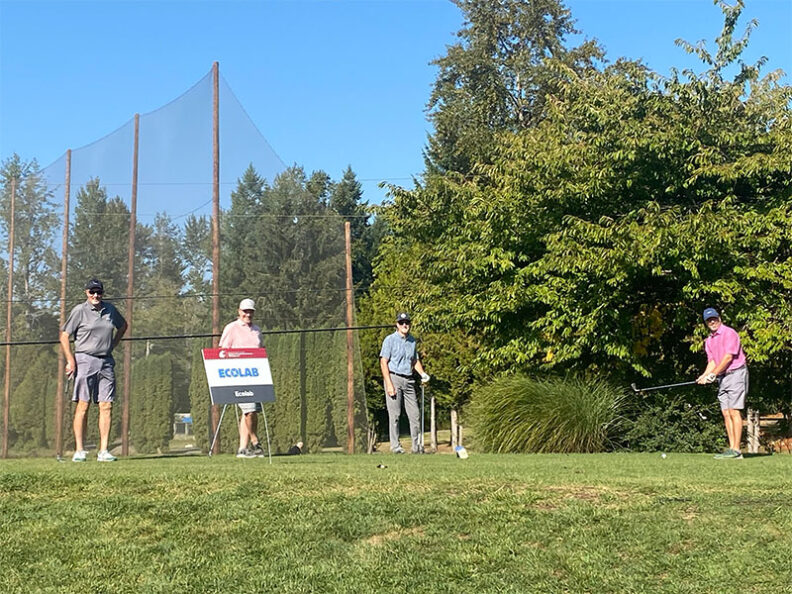 Four golfers standing on a tee box with clubs, next to an Ecolab sponsor sign, surrounded by green grass, trees, and a tall netting backdrop during the SHBM First Annual Golf Tournament.