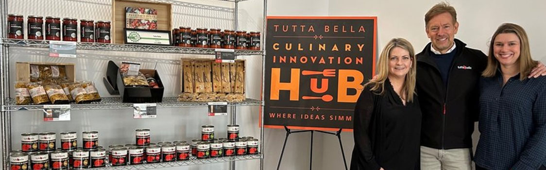 Shelving stocked with Tutta Bella branded sauces and packaged foods next to a sign reading “Tutta Bella Culinary Innovation Hub – Where Ideas Simmer,” with three individuals standing together (L to R: Kelly Parriott, VP of retail marketing & Innovation; Joe Fugere, Tutta Bella founder & CEO; Tamra Nelson, VP of retail sales).