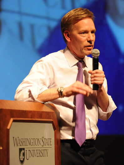 Joe Fugere speaking at a podium with a microphone during the Burtenshaw Lecture at Washington State University in 2011, with a purple tie and presentation screen in the background.