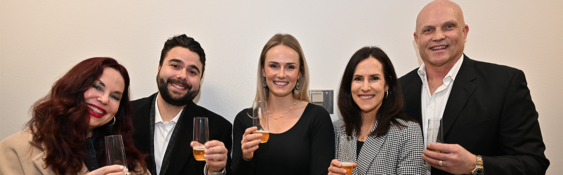 Five people standing together indoors holding champagne flutes filled with a golden beverage, dressed in business and semi-formal attire.