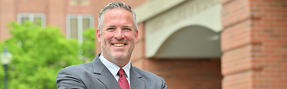 Jim Harbour wearing a gray suit with a red tie, standing outdoors in front of a brick building with an arched entrance and greenery in the background.