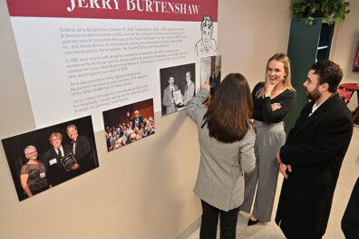Three Burtenshaw family members stand near a wall display honoring Jerry Burtenshaw, reading and discussing the signage that includes text and historical photos.