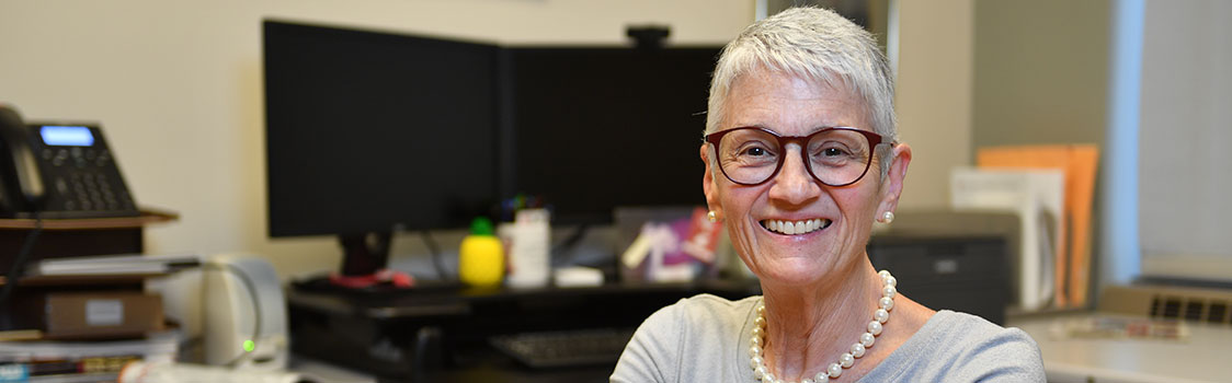 Nancy Swanger seated in an office environment with dual computer monitors, a desk phone, and assorted office supplies visible in the background.
