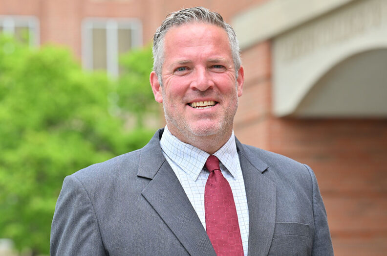 Jim Harbour wearing a gray suit with a red tie, standing outdoors in front of a brick building with an arched entrance and greenery in the background.