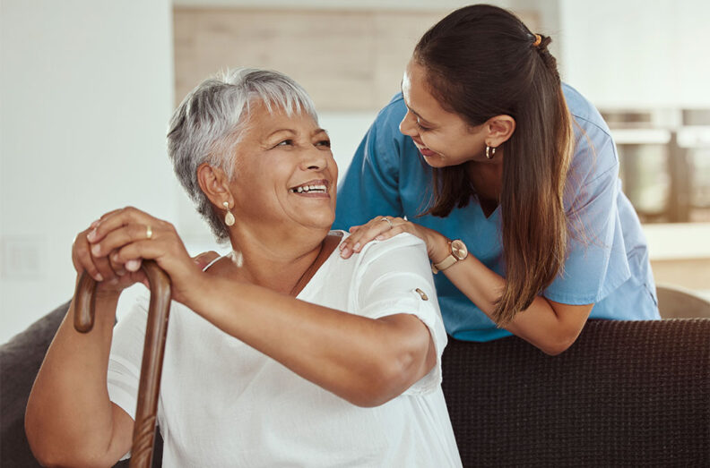 A person in a white shirt holding a wooden cane sits while another person in scrubs gently places hands on their shoulders in a supportive gesture.