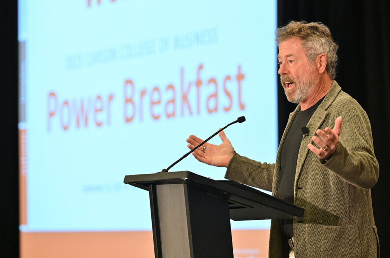 Kurt Beecher Dammeier stands at a podium gesturing with both hands during a presentation at the 2025 Power Breakfast, with a large screen behind displaying the event name.