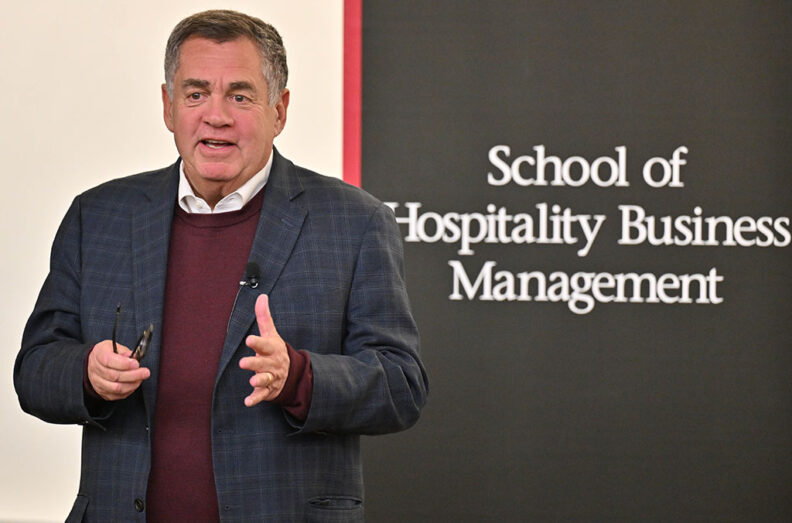 John Oppenheimer giving a presentation in front of a wall with repeated WSU branding and a black panel displaying the text “School of Hospitality Business Management.” The individual is wearing a dark plaid jacket over a maroon sweater and holding a presentation remote.