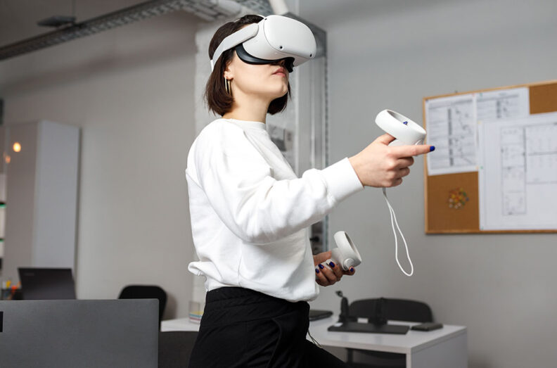Person wearing a white sweatshirt using a virtual reality headset and handheld controllers in an office setting with a corkboard and papers in the background.
